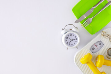 Proper nutrition concept. Flat lay composition made of scales, plate with cutlery, dumbbells, tape measure, alarm clock on white background with copy space.