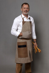 A man in a linen kitchen apron with potholders holds kitchen utensils.