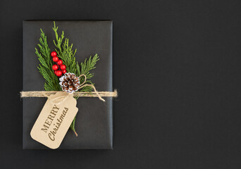 Black gift box with craft label with the text merry christmas, cone, twigs of cedar tree and berries on a dark background. Space for yout text.