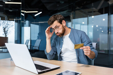 Cheated and disappointed businessman working inside office, man holding bank credit card and trying...