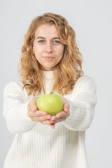 A young pretty curly blonde woman holds a yellow apple in her hands. The girl looks happy and smiles, she is dressed in a white knitted suit. Near the model there is a lot of space for a