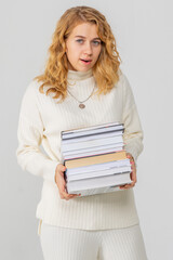 A young attractive blonde woman holds a stack of books in her hands. The girl is dressed in a knitted white sweater. The student rejoices at the passed session and smiles