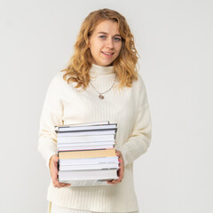 A young attractive blonde woman holds a stack of books in her hands. The girl is dressed in a knitted white sweater. The student rejoices at the passed session and smiles