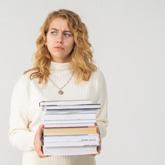 A young attractive blonde woman holds a stack of books in her hands. The girl is dressed in a knitted white sweater. The student does not want to study and looks dissatisfied