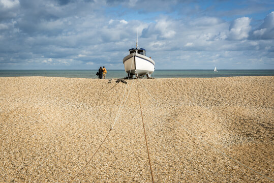 Couple stands next to a tethered cabin boat on a shingle beach, Dungeness, Kent, England, UK