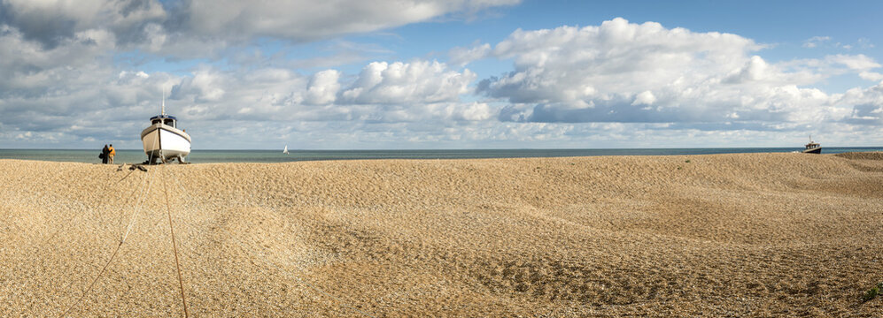 Couple stands at the water's edge looking out into the ocean next to a wooden cabin boat tethered on the shingle beach at Dungeness along the Atlantic Coast; Dungeness, Kent, England, United Kingdom