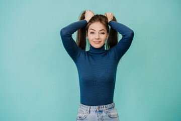 Young adult pretty woman in blue sweater, jeans looks at camera, holding her hair, making two...