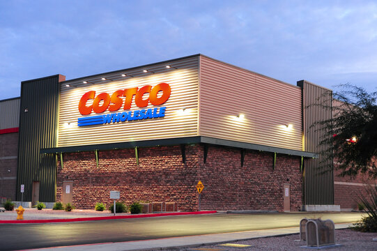 An exterior view of a Costco Wholesale store in Arizona, showcasing its well-lit sign on a brick and metal facade. The building&rsquo;s design and iconic branding stand out against the evening sky