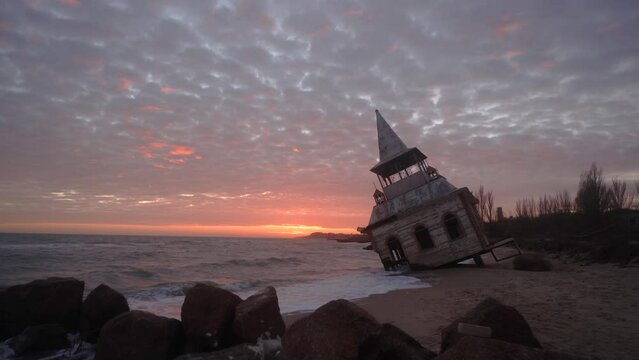 Mysterious vintage flooded leaning wooden house or chapel washed by sea waves, at coastline on pink sunset. Enigmatic rickety building sinking in water on sandy beach near breakwater on sunrise.