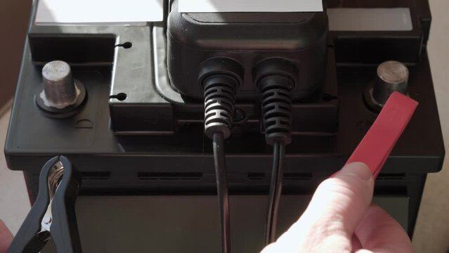 Recharging a car battery with a charger. A man's hand puts clamps on the battery terminals.