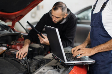 Fototapeta na wymiar Close up view of hand of young male mechanic using laptop and his colleague with tablet, recording automobile engine checking collecting detailed information during their work on car workshop.