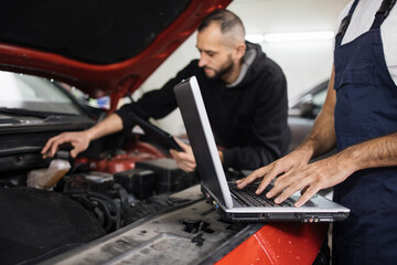 Close up view of hand of young male mechanic using laptop and his colleague with tablet, recording automobile engine checking collecting detailed information during his work on car workshop.