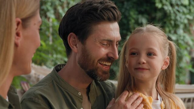 Close up Caucasian adult parents bearded father dad and mother talking funny smile laugh speak happy conversation with small cute child girl daughter daddy cuddle hug baby laughing together outdoors