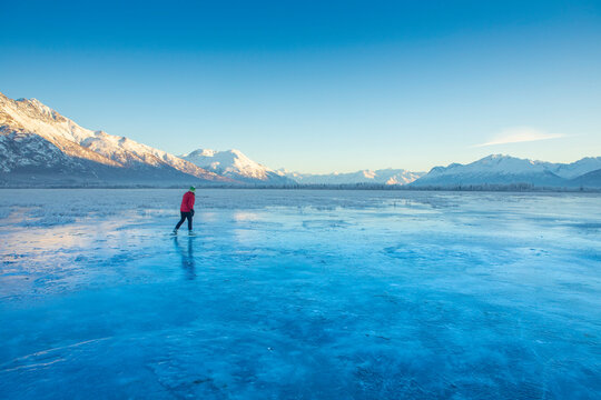 A caucasian woman backcountry ice skating, nordic blading on Gull Lake in Palmer, on a winter day with the sun lighting up the Chugach Mountains in the background; Alaska, United States of America