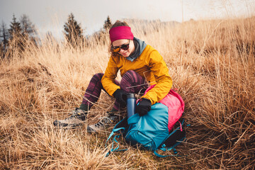 Woman hiker sitting on the ground opening her backpack, taking out a blue thermo flask