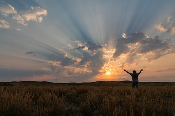 Corpuscular rays during sunrise over the prairies of Saskatchewan. Woman standing and rejoicing in the beauty of a new day; Saskatchewan, Canada