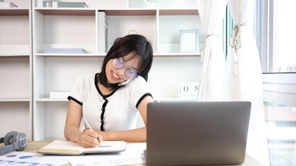 Asian female worker talking on phone and working on laptop with smiling face, Office staff work and telephone conversations, Take notes for reminders, Business conversation over the phone.