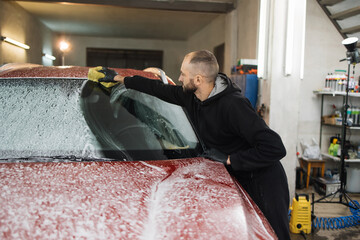 Confident male car wash worker cleaning the windshield of red luxury car with help of special foam...