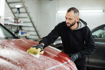 Young bearded car service male worker doing cleaning the hood of red crossover car with the help of...