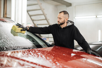 Car wash and clean with shampoo and sponge. Young handsome bearded man worker washing soapy car...