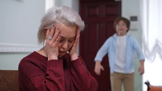 Portrait of devastated Caucasian mature woman thinking as blurred schoolboy yelling misbehaving at background. Sad stressed mother with disobedient son indoors