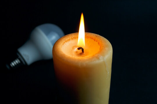 Burning candle and switched off light bulb in darkness. Blackout, electricity off, energy crisis or power outage, concept image.