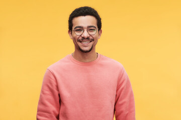Young cheerful man in eyeglasses and pink sweatshirt looking at camera while standing against vivid yellow background in isolation - obrazy, fototapety, plakaty