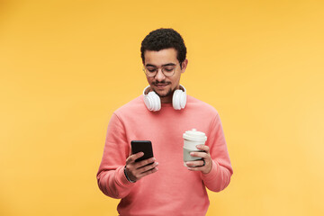 Young multi-ethnic man with white headphones around neck using smartphone and having cup of coffee...