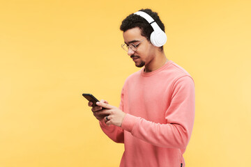 Young man in headphones and pink pullover scrolling through playlist in mobile phone while standing in front of camera on yellow background