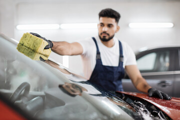 Young bearded man, car wash worker, wearing t-shirt and overalls cleaning the windshield of car with the help of special foam and sponge. Selective focus on yellow sponge.
