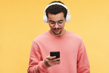 Young man in pink pullover listening to music in headphones and choosing new sound tracks in mobile phone in front of camera