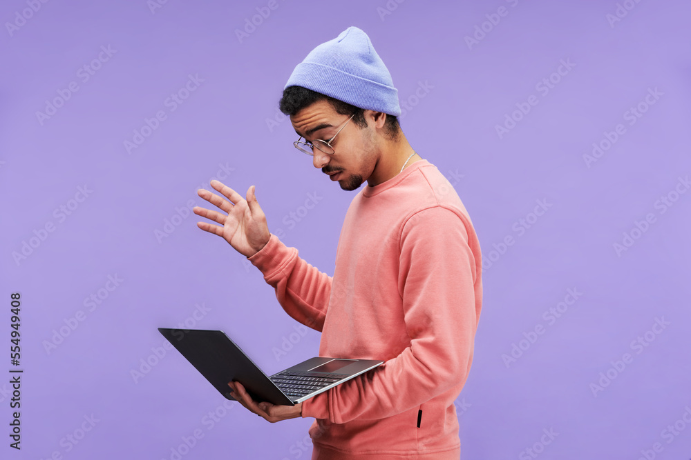 Poster side view of young astonished male programmer in casualwear and eyeglasses looking at laptop screen 