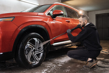 Car wheel, rim or alloy, washing. Car cleaning with water jet. Horizontal shot of pleasant young guy in white t-shirt and overalls, washing red car wheel with high pressure water jet.