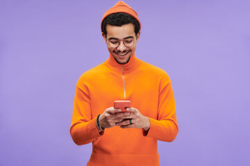 Smiling multi-ethnic guy in orange casualwear texting in smartphone or reading advert message or...
