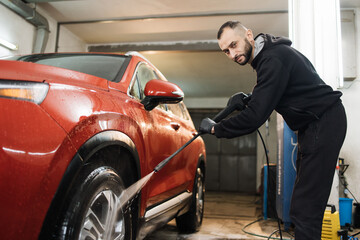 Washing car with high pressure water at car wash service. Close up of red car wheel indoor. Smiling bearded man washing red car with water jet.