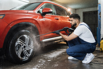 Side view portrait of young bearded male worker washing the car wheels rims with a high pressure...