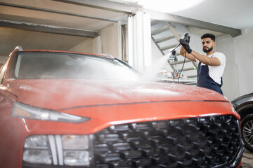 Cleaning car using high pressure water gun. Handsome young bearded man worker washing modern red luxury car under high pressure water in professional car wash service.