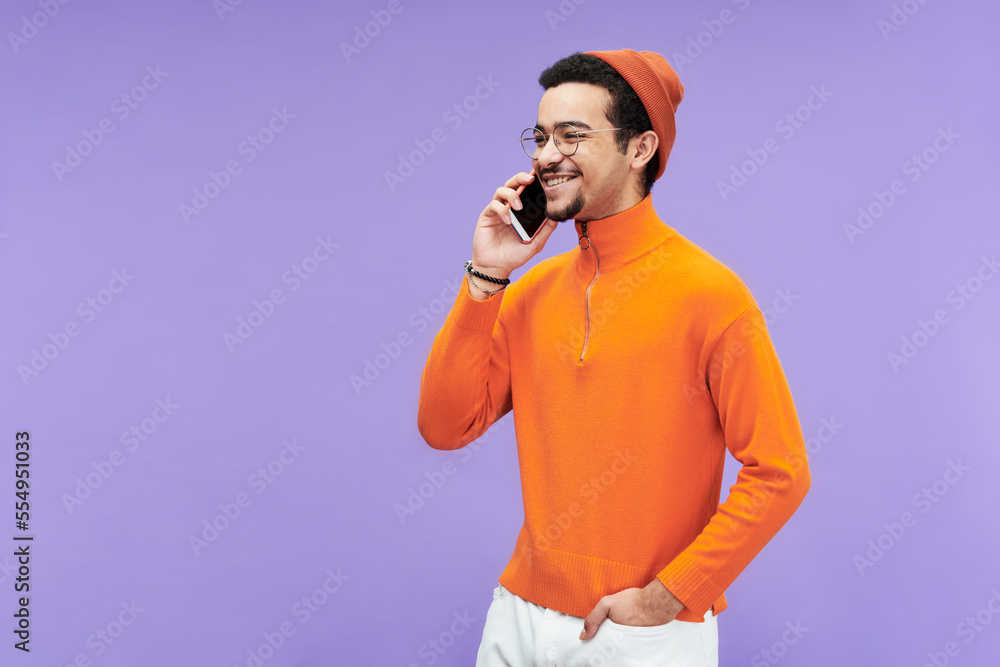 Poster happy young man in turtleneck, beanie cap and eyeglasses communicating on smartphone while standing 