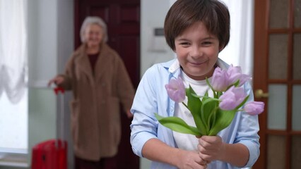 Charming Caucasian son rejoicing standing on the right with bouquet of flowers looking back at blurred mother returning home. Portrait of happy smiling boy looking at camera posing indoors