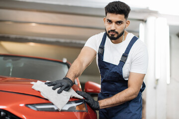 Low angle view of male professional car wash worker in black rubber gloves, holding the gray microfiber and polishing the car hood and headlight of luxury red car.
