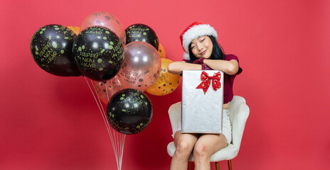 Happy young woman with santa hat and big giftbox sitting comfort with balloons happy new year. over red background.  Concept of the New Year and Christmas Day.