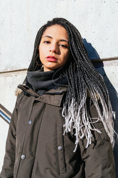 Young stylish female with long black braids and in outerwear leaning on wall in city and looking at camera
