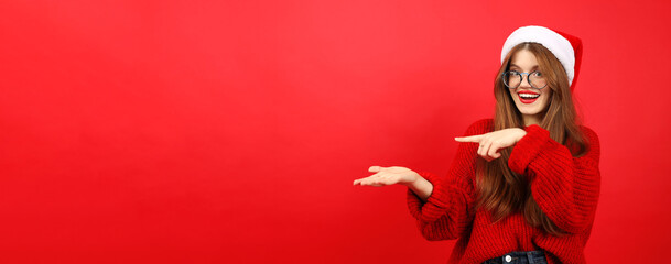 A young woman in a Santa hat and a Christmas sweater points to an empty space in her hands for text...