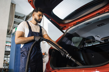Bearded male worker wearing white t-shirt and blue overalls, vacuuming car interior, trunk with wet vacuum cleaner, professionally extraction method. Wet car vacuum cleaning.