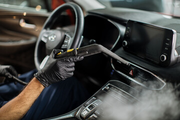 Plakat Close up of hands of man in black protective rubber gloves cleaning interior of the car with hot steam cleaner. Selective focus on guy hands. Auto cleaning service and detailing concept.