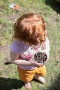 A red headed toddler playing with a pot of earth in a garden by herself.