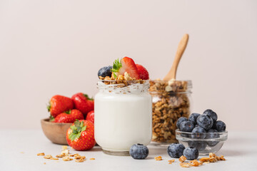 Natural yogurt in jar with granola and fresh strawberries and blueberries on light table and beige...
