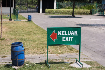View of green metal information board with keluar (exit) red direction arrow in public park with blue plastic trash bin and grass and trees. No people.