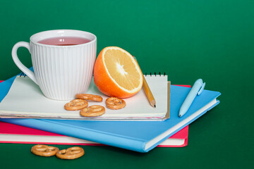 Notebooks, a cup of tea and an orange on a green background, copy space.