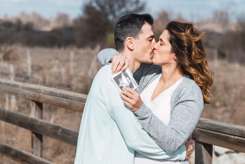 Couple kissing outdoors, she is pregnant and holding a sonogram in her hands